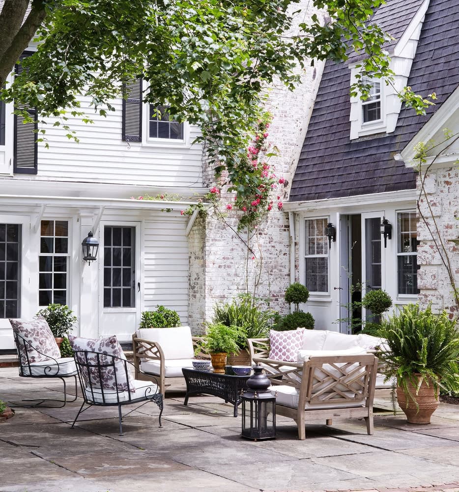 A cozy raised deck patio with cushioned wooden and metal chairs, potted plants, and a coffee table, set in front of a white house with dark roof shingles and climbing flowers on the wall.
