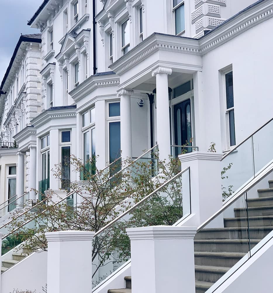 Row of elegant white Victorian-style terraced houses with large windows, decorative columns, and glass-railed staircases leading up to the entrances—perfect inspiration for those seeking home remodeling tips. Small trees are planted beside the stairs.