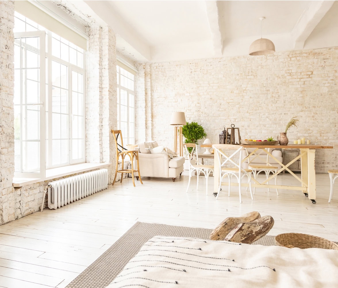 A bright, airy living and dining area by a general contractor in Northern VA, featuring white brick walls, large windows, light wooden floors, white chairs, a beige sofa, and a wooden dining table decorated with plants and candles.