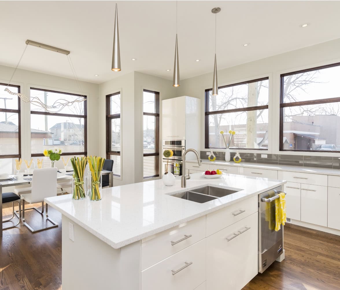 Bright, modern kitchen with white cabinets, an island sink, stainless steel appliances, pendant lights, and large windows. Designed by a leading general contractor in Northern VA, this space features yellow accents and a stylish dining area with white chairs.