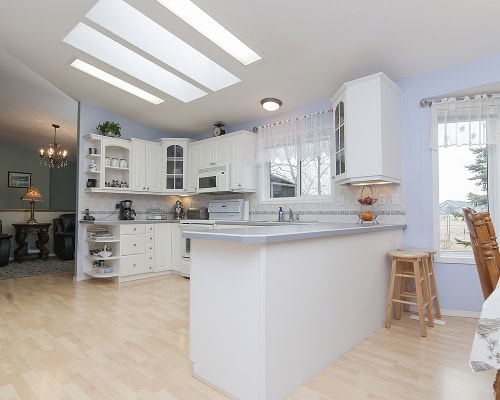 Bright, modern kitchen with white cabinets, light wood flooring, skylights, and a breakfast bar with two wooden stools. Designed by a skilled kitchen remodeling contractor, with natural light and views of the adjacent dining and living area.