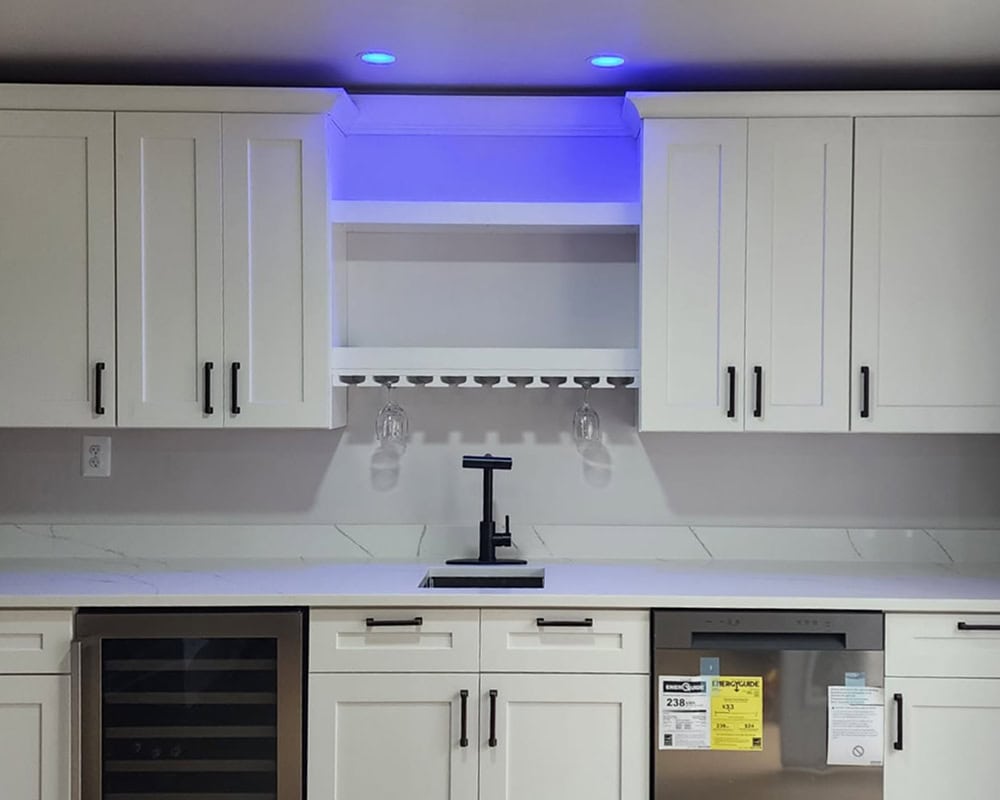 Modern kitchen with white cabinets, a built-in wine glass rack, two hanging wine glasses, under-cabinet blue LED lighting, black faucet, marble countertop, and stainless steel dishwasher—perfectly crafted by a skilled kitchen remodeling contractor.