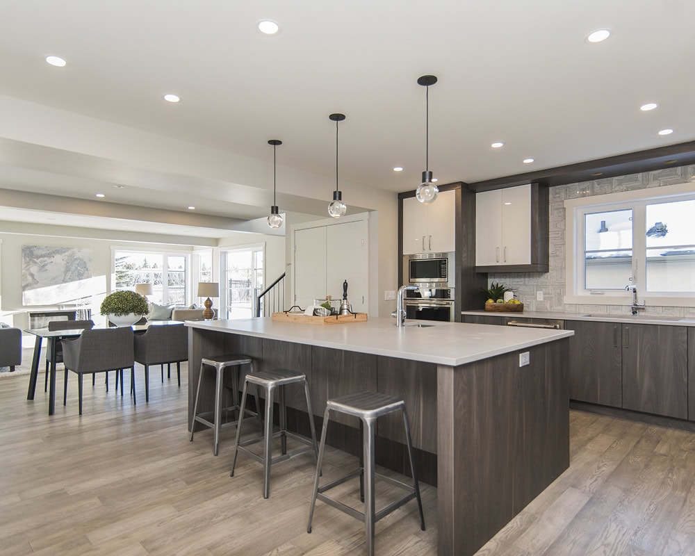 Modern kitchen with gray wood cabinetry, a large island with three metal stools, pendant lighting, stainless steel appliances, and an adjacent dining area—expertly crafted by a skilled kitchen remodeling contractor.