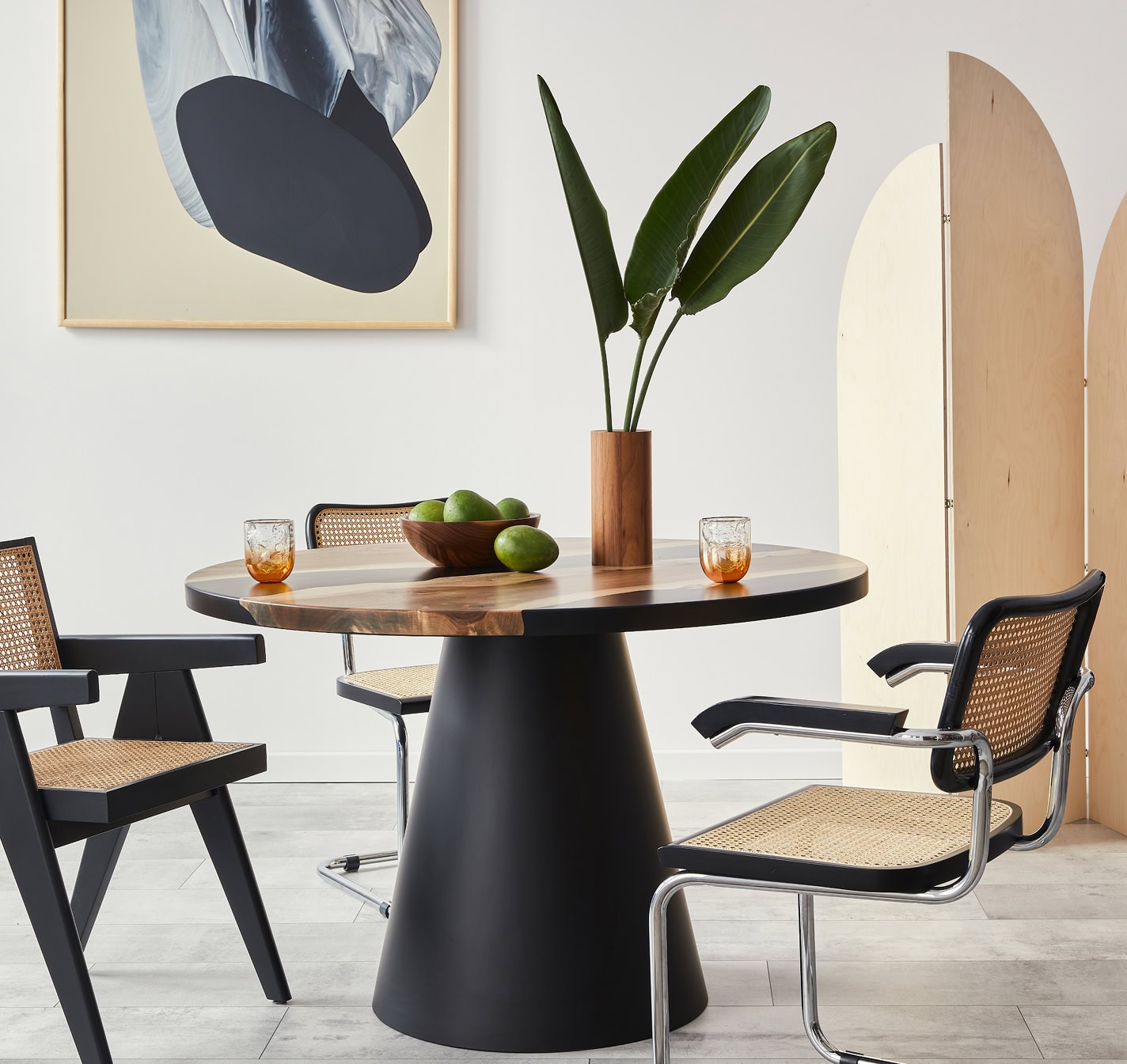 A modern dining area by a residential remodeling company features a round wooden table, two woven chairs, a vase with tall green leaves, a bowl of limes, and two glasses. Abstract art and a light wood divider decorate the background.